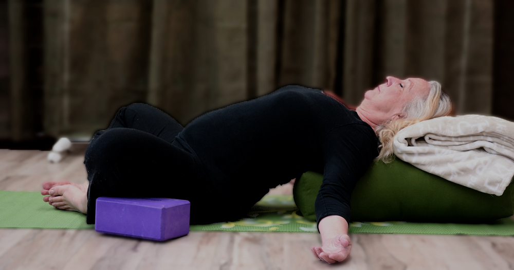 Woman lying on back on yoga mat and bolster, eyes closed.